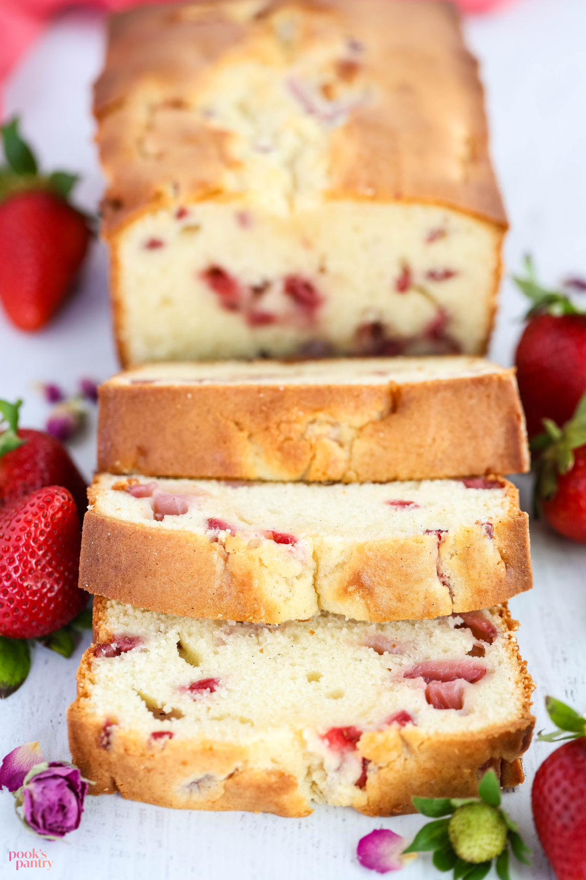 Close up photo of slices of strawberry cream cheese poundcake with fresh strawberries and dried rose buds scattered around.