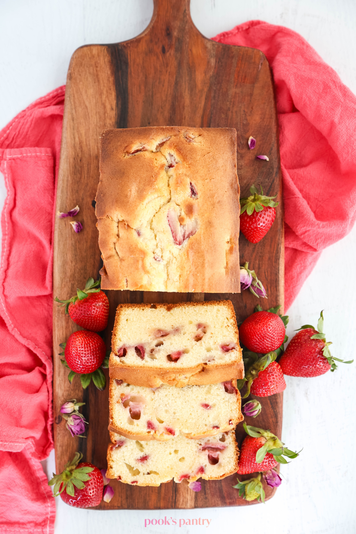 Overhead photo of sliced cake with strawberries on a wooden cutting board, sitting on a pink tea towel.