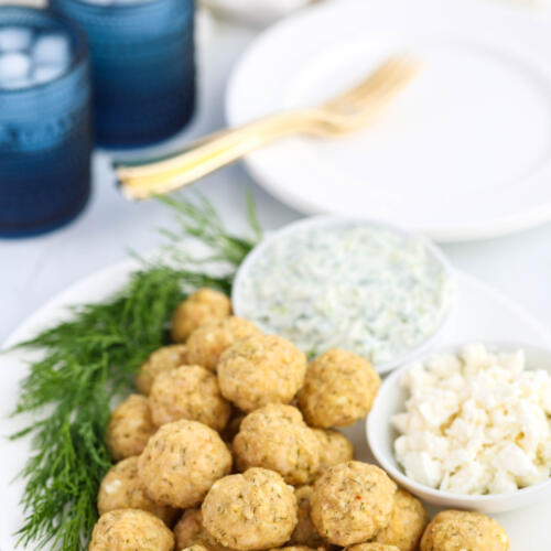 Baked Mediterranean chicken meatballs on a white plate with small bowls of feta and tzatziki sauce, with fresh dill on the plate.