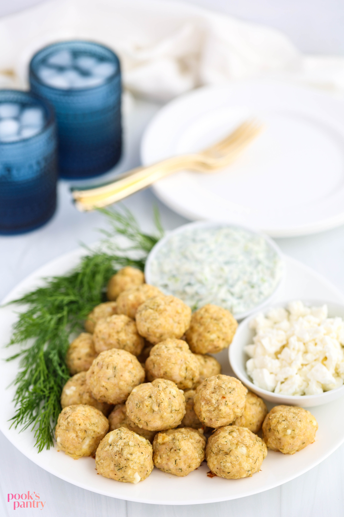 Baked Mediterranean chicken meatballs on a white plate with small bowls of feta and tzatziki sauce, with fresh dill on the plate.