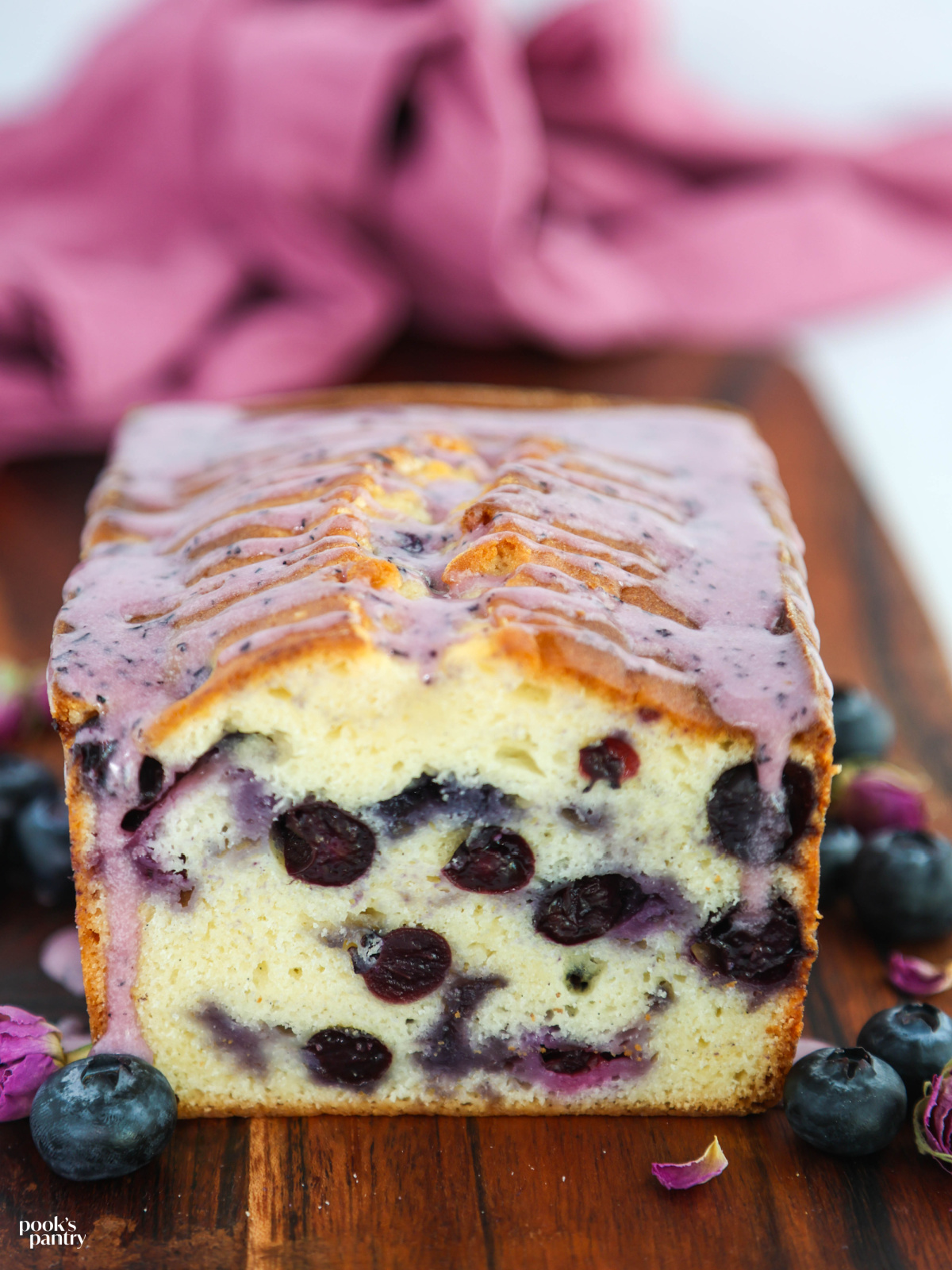 Pound cake recipe with a fresh blueberry glazed drizzled on top. The cake is on a wooden board with dried flowers and fresh berries on the sides.