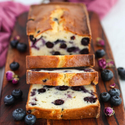 Moist blueberry pound cake recipe on a wooden board with fresh blueberries and dried rosebuds on the sides.