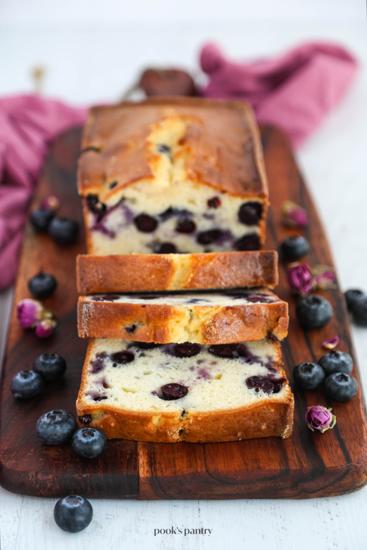 Moist blueberry pound cake recipe on a wooden board with fresh blueberries and dried rosebuds on the sides.