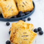 Blueberry mini pies on a white background with a plate of pies and blueberries in the background.