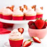 Vanilla cupcake with strawberry cream cheese frosting garnished with a candied strawberry slice on a small white plate in front of a platter of cupcakes.