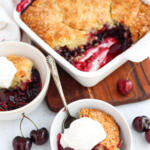 Bowls of cherry cobbler with whipped cream on top, with a dish of cobbler on a wooden board in the background.