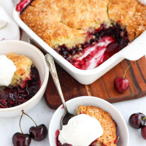 Bowls of cherry cobbler with whipped cream on top, with a dish of cobbler on a wooden board in the background.