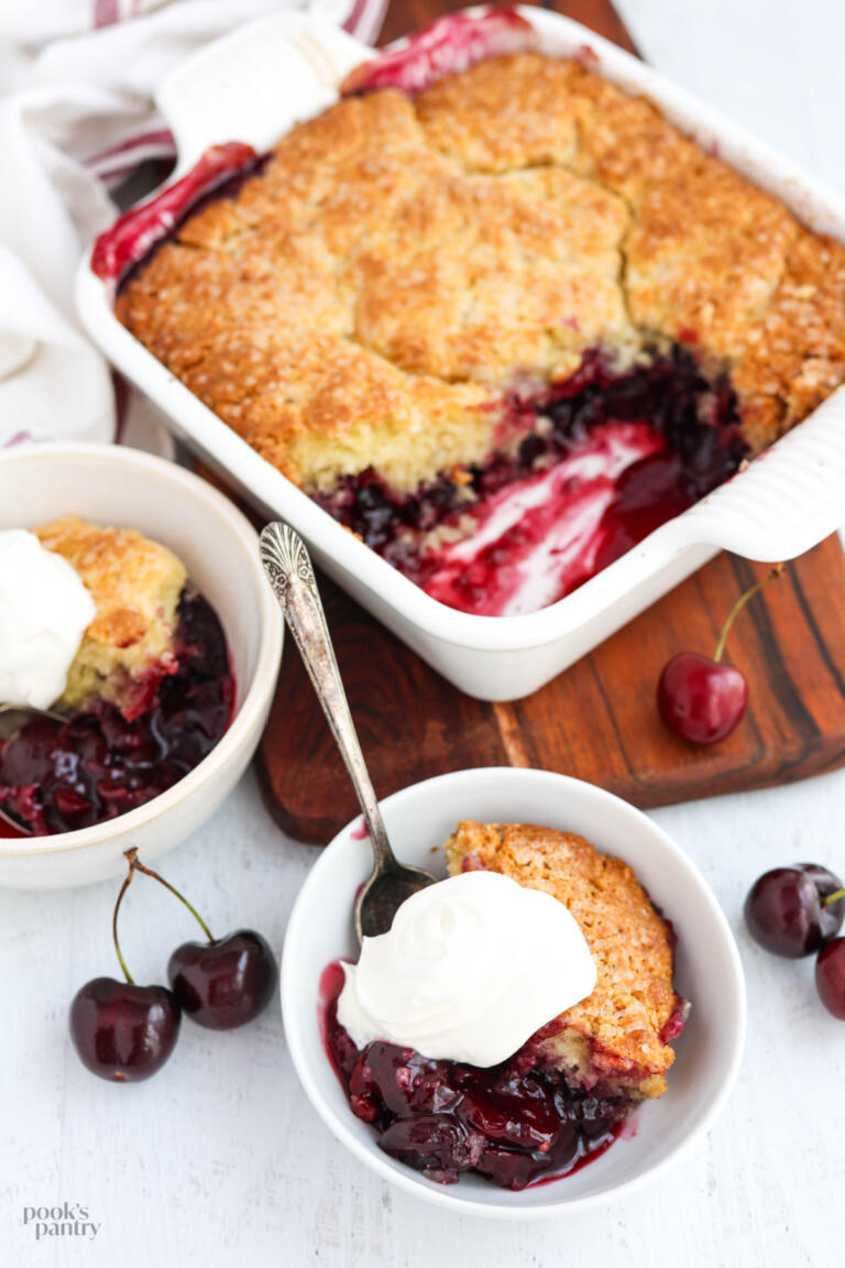 Bowls of cherry cobbler with whipped cream on top, with a dish of cobbler on a wooden board in the background.