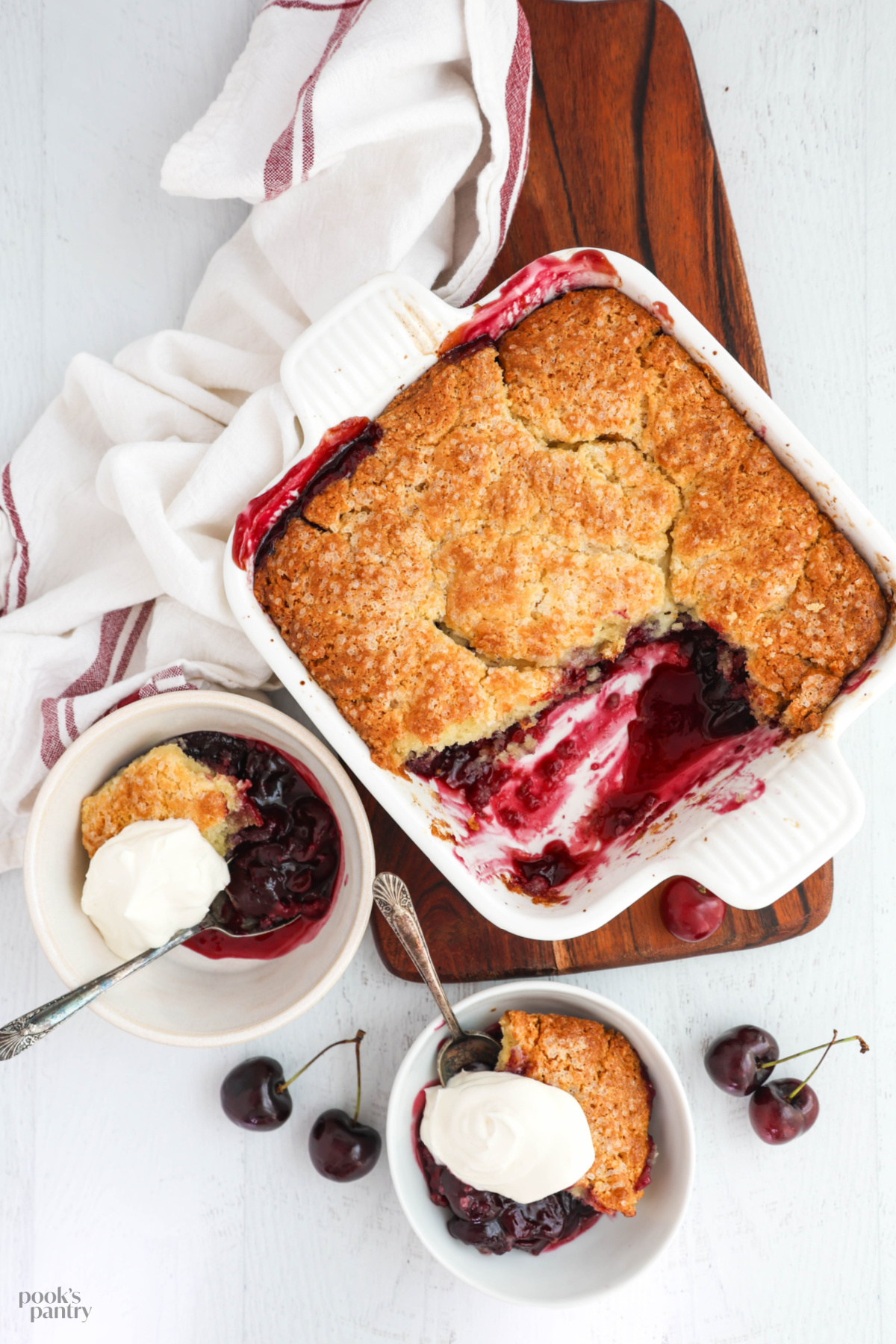 Overhead photo of a baking dish and two bowls of cherry cobbler, sitting on a walnut cutting board with a tea towel on the side.
