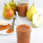 Pear butter in a canning jar with a silver spoon in it. There are pears in the background, with another jar of pear butter in the middle.