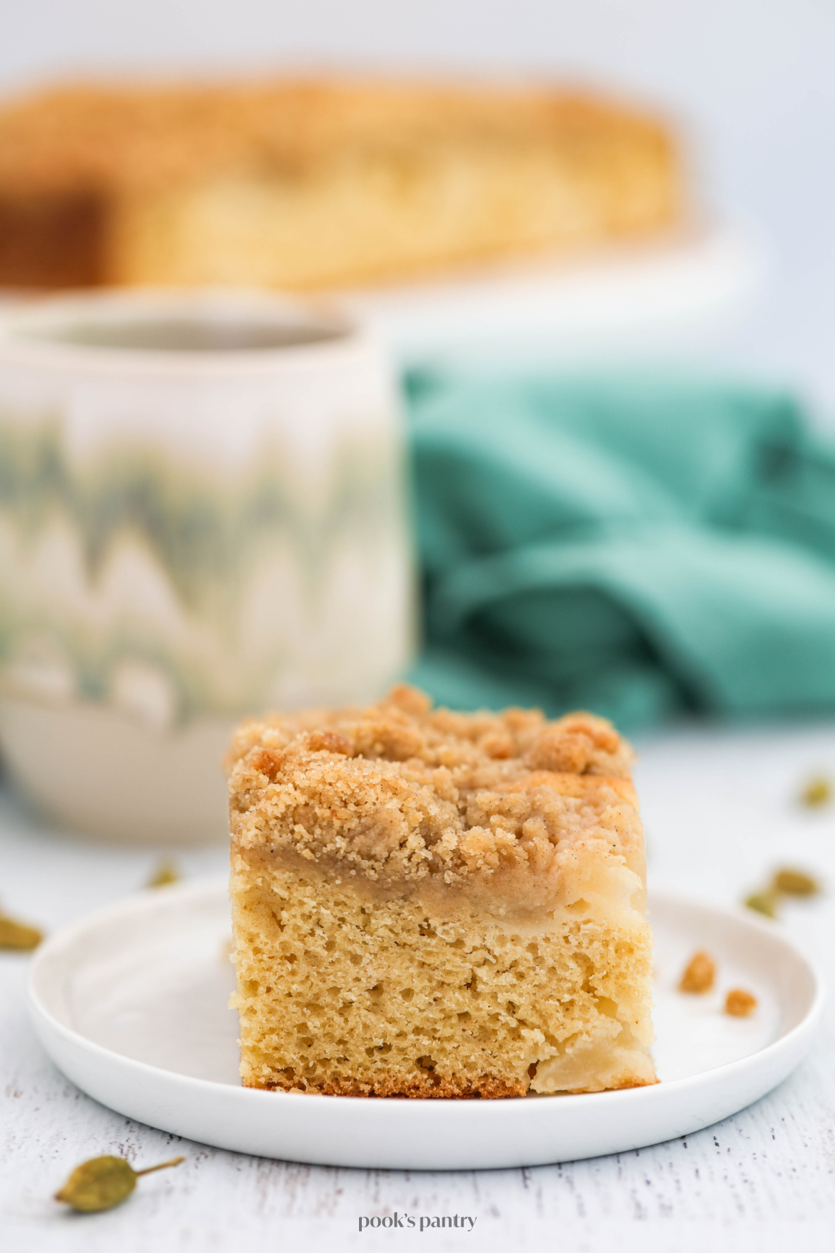 A slice of coffee cake on a small white plate with a cardamom pod in front of the plate. There is a mug behind the plate and a kitchen towel.