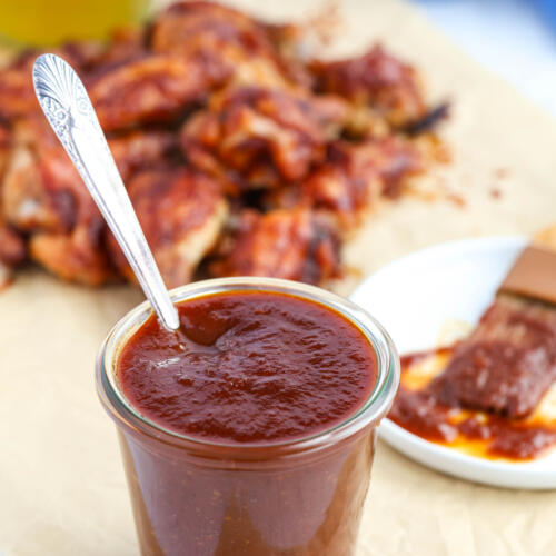 Maple bourbon BBQ sauce in a clear glass jar with an antique spoon in the jar. There are sauced wings in the background.