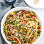 Roasted vegetable pasta topped with herbs and grated parmesan in a wide white bowl. There are blue glasses filled with ice water in the top left corner.