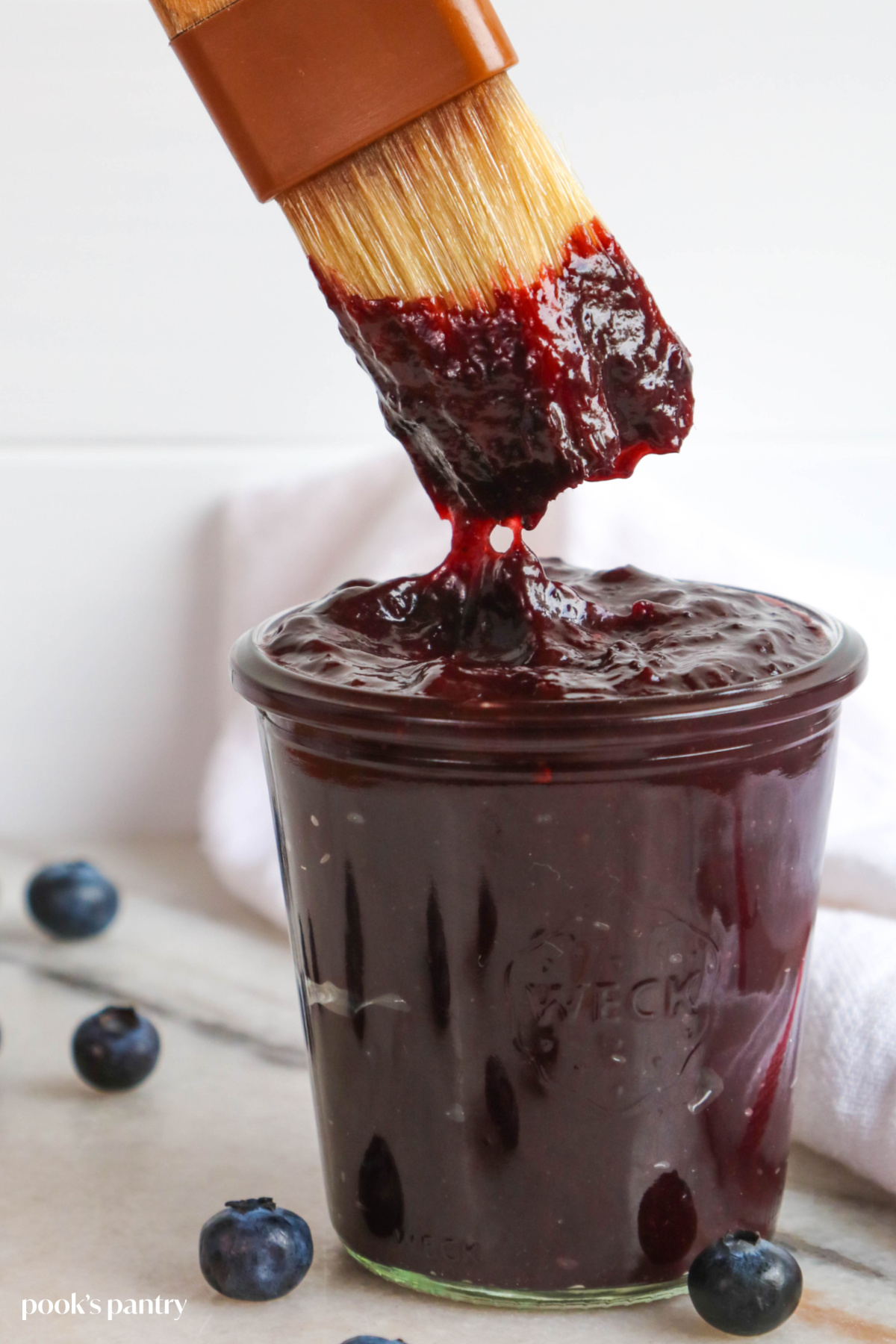 Dipping a brush into a jar of blueberry bourbon BBQ sauce. There are blueberries scattered around the jar.