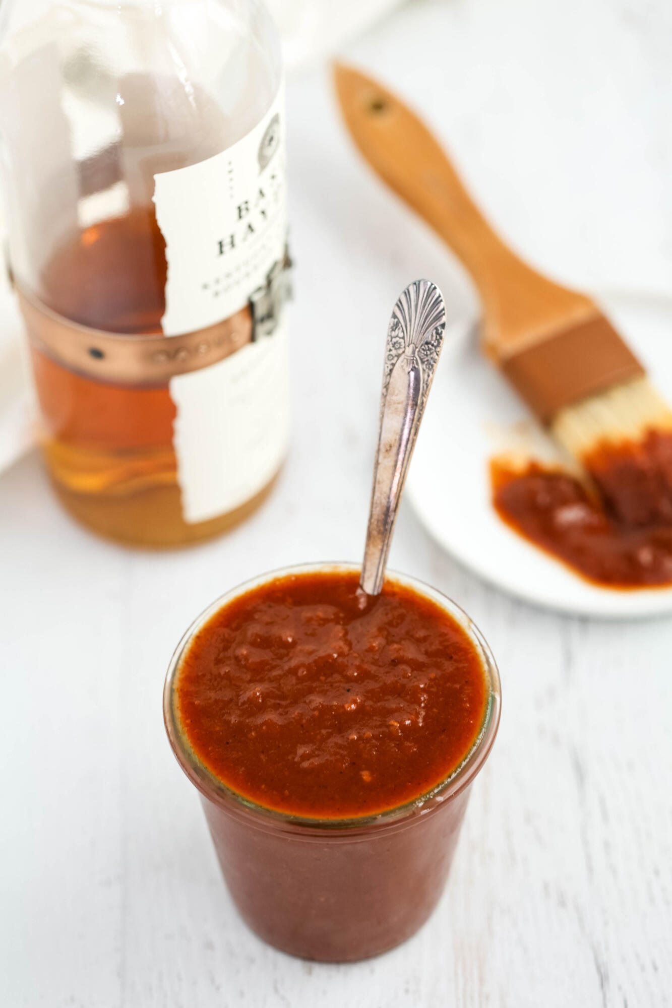 Maple bourbon barbecue sauce in a glass jar, with a bottle of bourbon in the background.