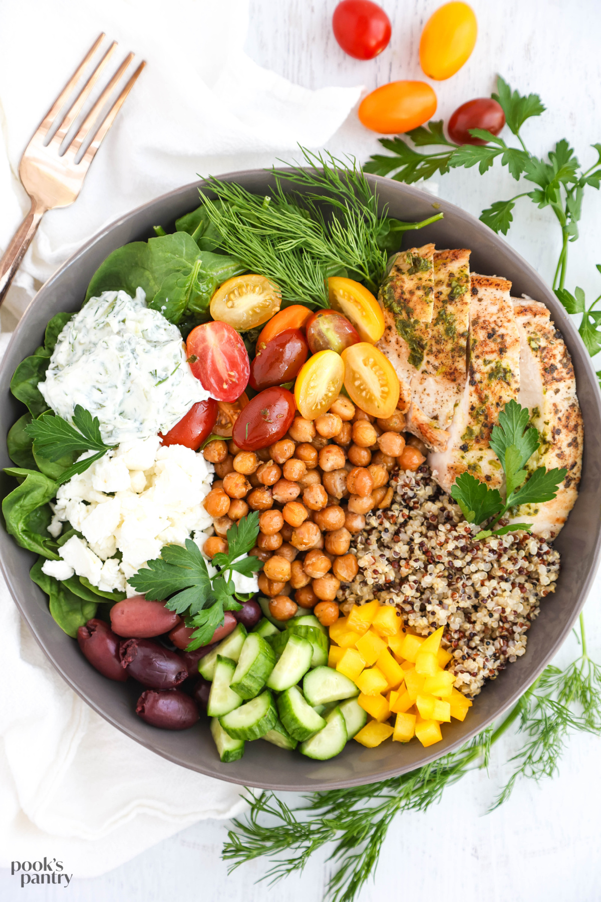 Mediterranean chicken meal prep with feta and quinoa in a gray bowl with herbs surrounding the bowl.