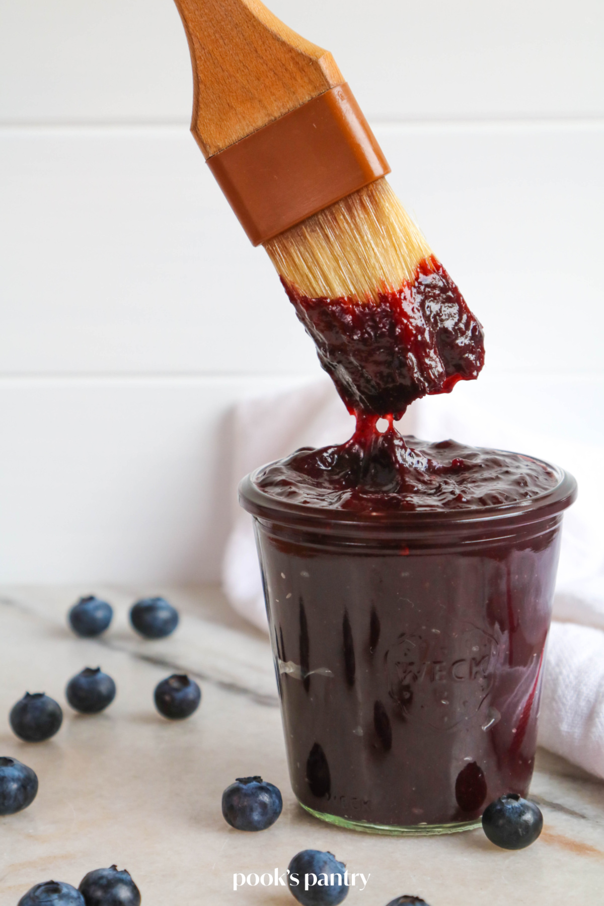Dunking a brush into a glass jar of BBQ sauce with blueberries.
There are blueberries scattered around the jar.