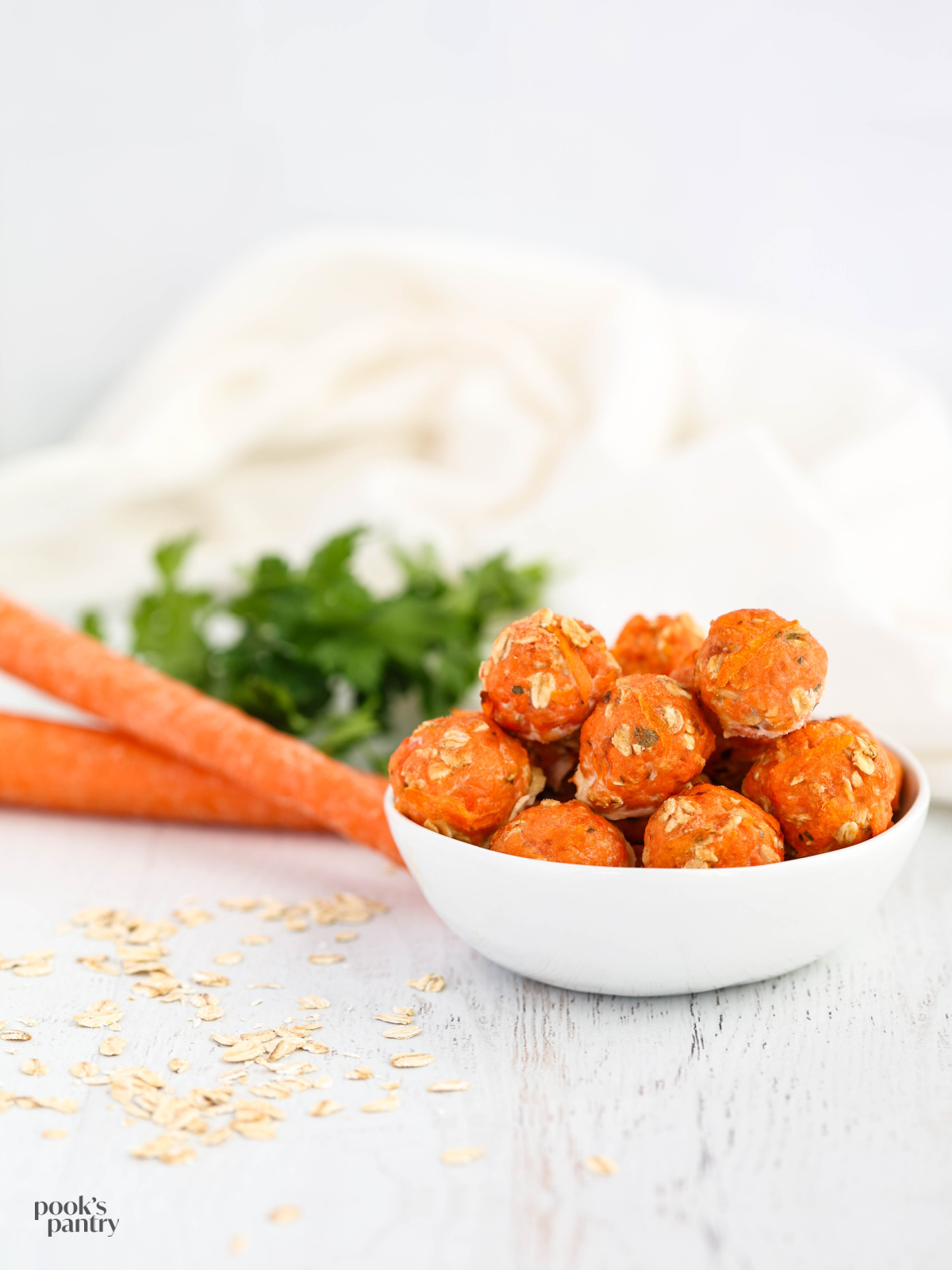 Bowl of salmon meatballs the right of carrots and parsley, with oats scattered around the bowl.