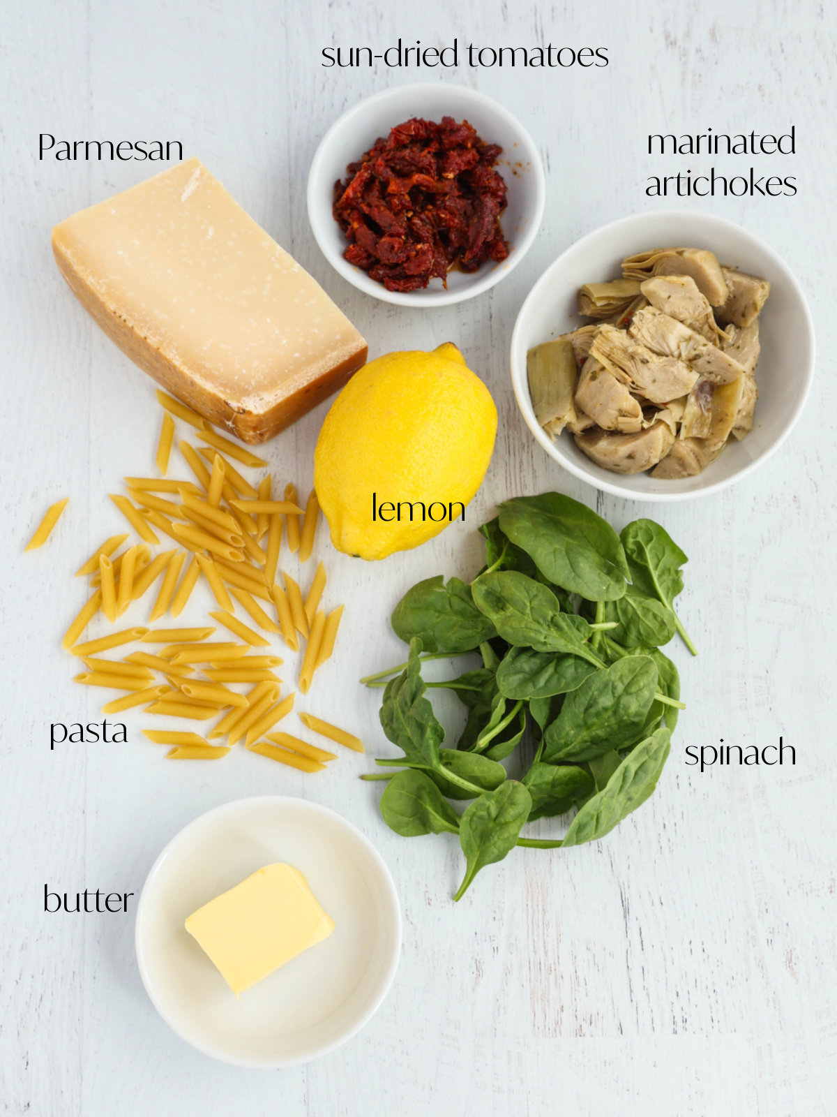 Ingredients for spinach artichoke pasta salad including pasta, spinach, artichokes, sun-dried tomatoes, lemon, and parmesan.