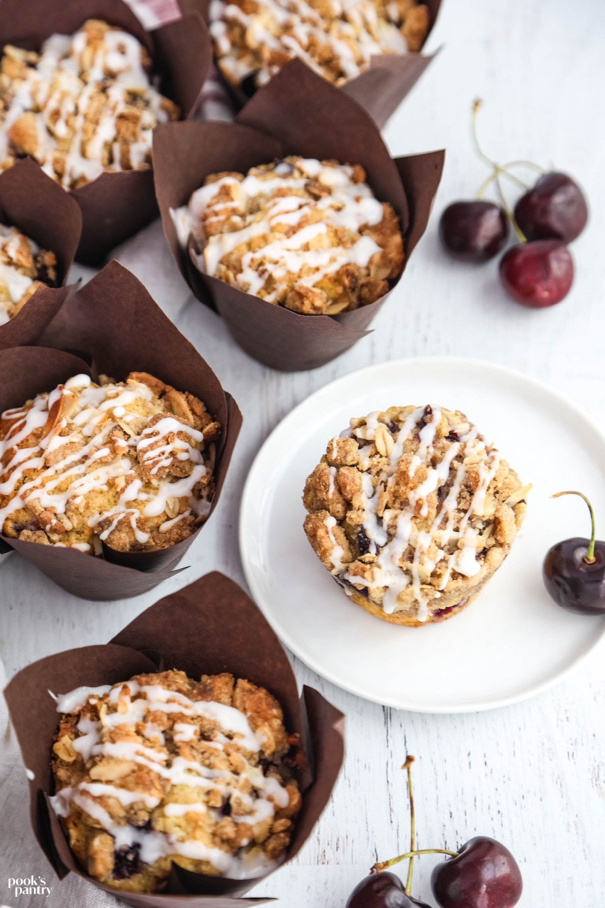 Homemade muffins in brown tulip liners with fresh cherries on the side. There is one muffin on a small plate to the right.