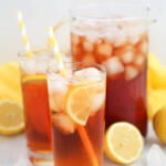 Two glasses of lemonade sun tea with a pitcher of tea in the background. There are yellow and white striped straws in the glasses.