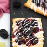 Blackberry puff pastry tarts on a black board with limes and blackberries on the side.