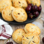 Three cherry hand pies stacked together in the forefront of the photo with a plate of hand pies in the background.