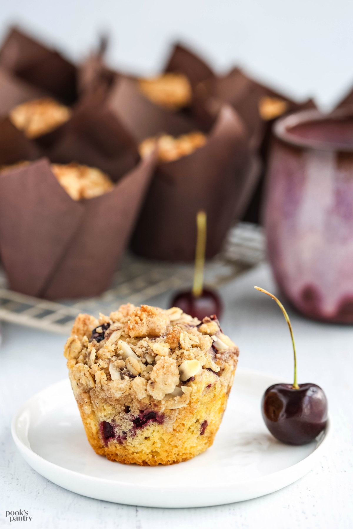 Cherry muffin on a small white plate with one cherry standing up beside it.