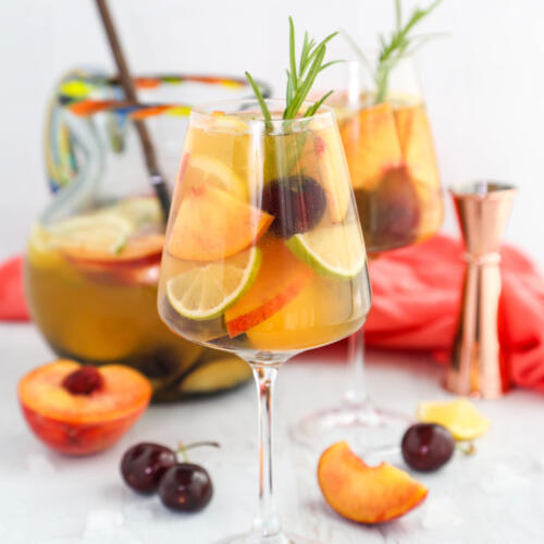 A glass of white peach sangria with a pitcher of sangria in the background.