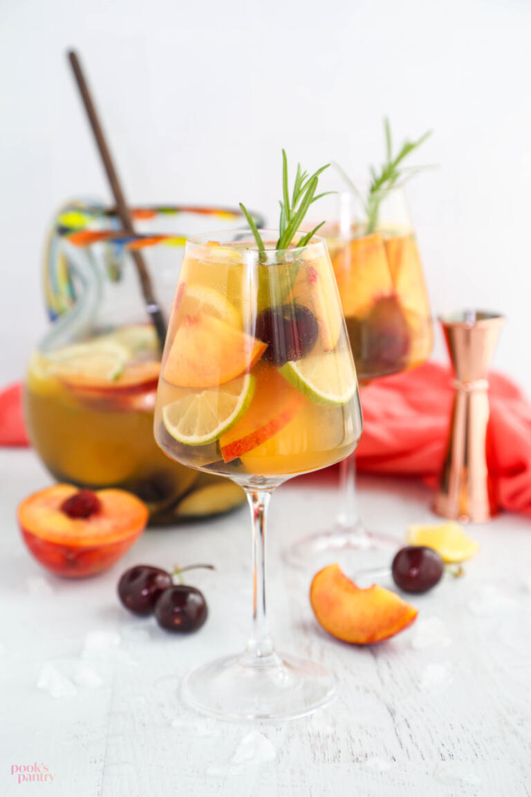 A glass of white peach sangria with a pitcher of sangria in the background.