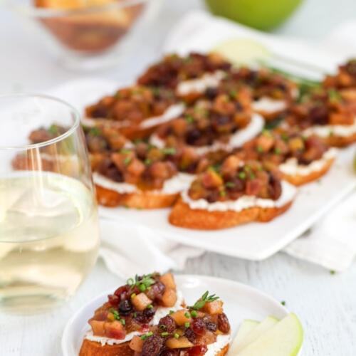 Two pieces of apple chutney bruschetta on a small white plate, with a platter of crostini in the background.