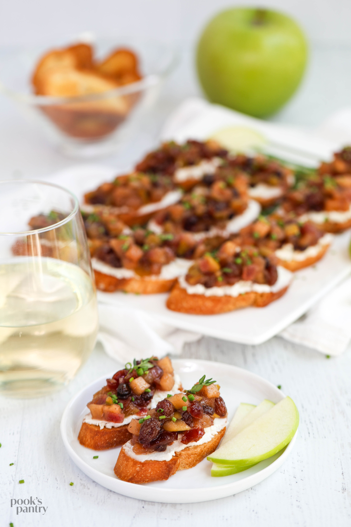 Two pieces of apple chutney bruschetta on a small white plate, with a platter of crostini in the background.