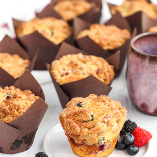 Bakery-style mixed berry muffins with crumble topping in brown paper liners, with one unwrapped muffin on a small plate.