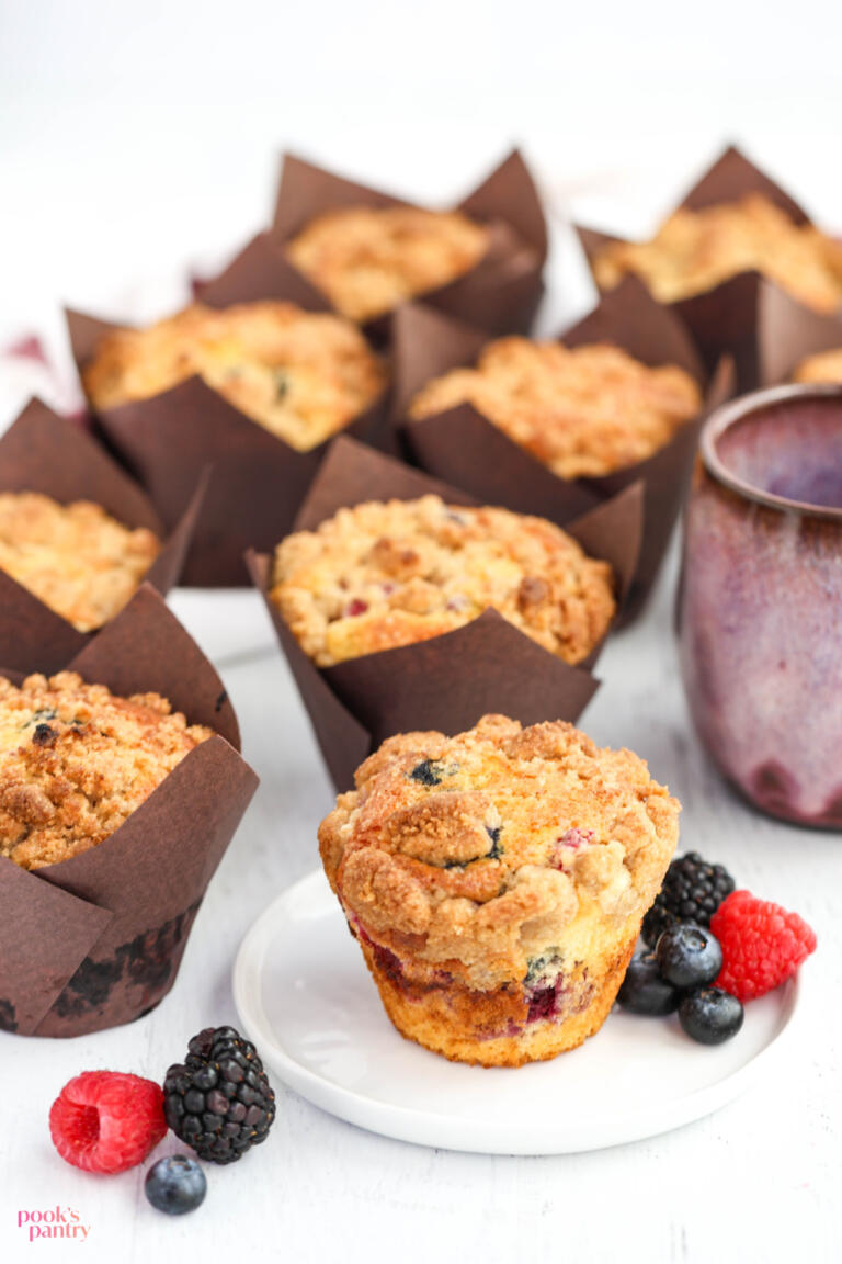 Bakery-style mixed berry muffins with crumble topping in brown paper liners, with one unwrapped muffin on a small plate.