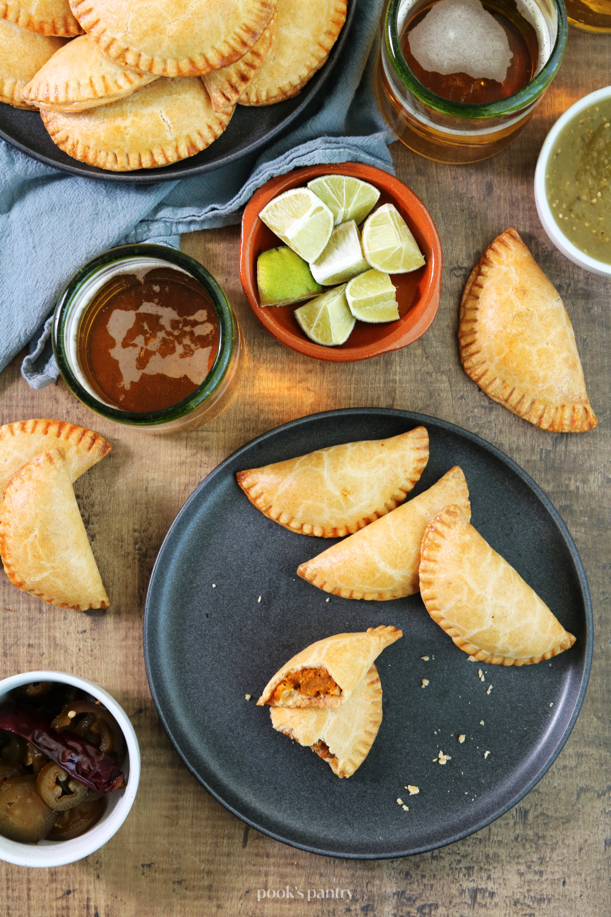 Savory pumpkin empanadas on a charcoal grey plate with glasses of beer, and dishes of salsa and cut limes in the background. A perfect fall appetizer for gatherings.