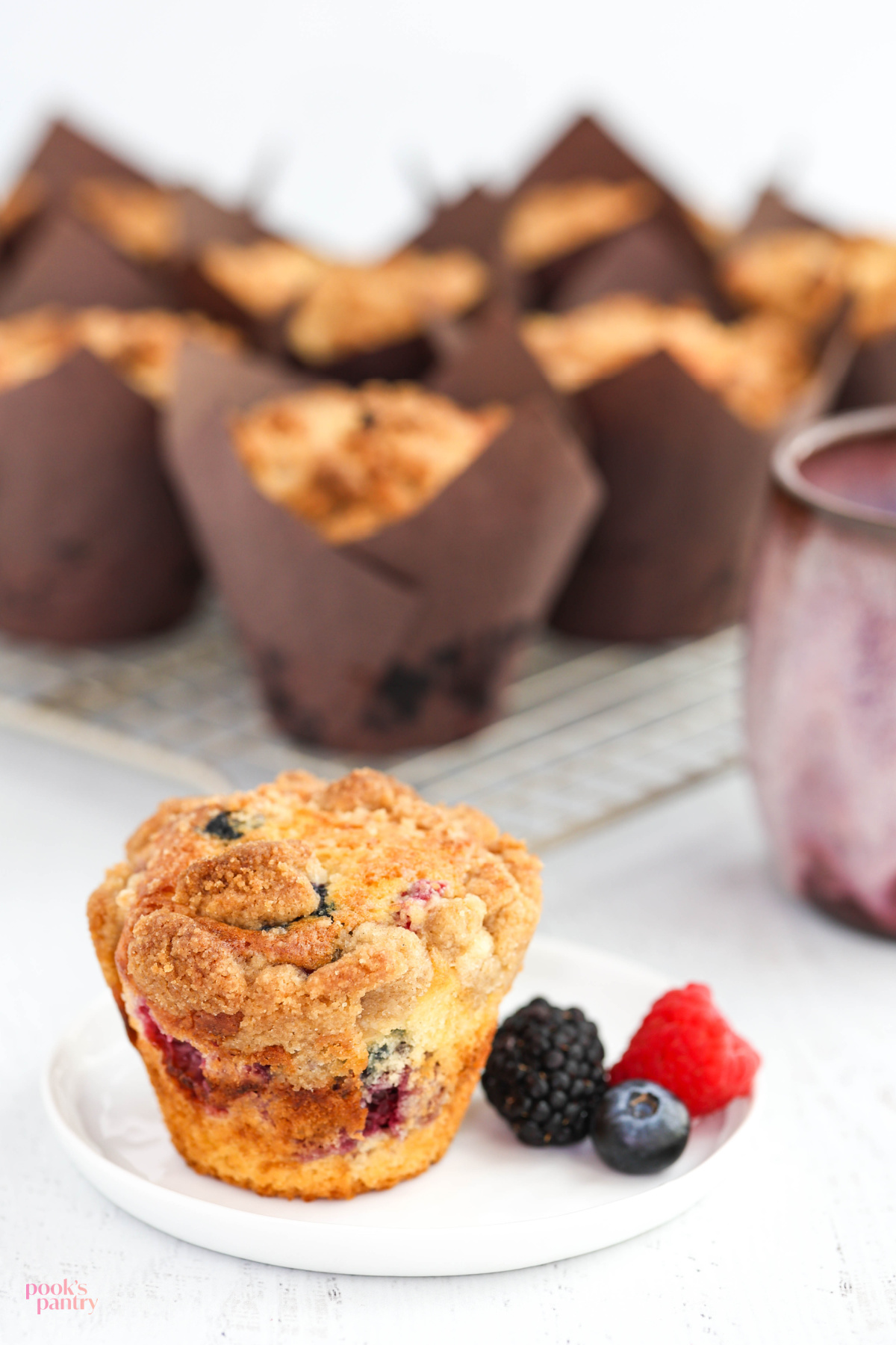 A triple-berry muffin sitting on a small white plate sitting in front of muffins on a cooling rack.