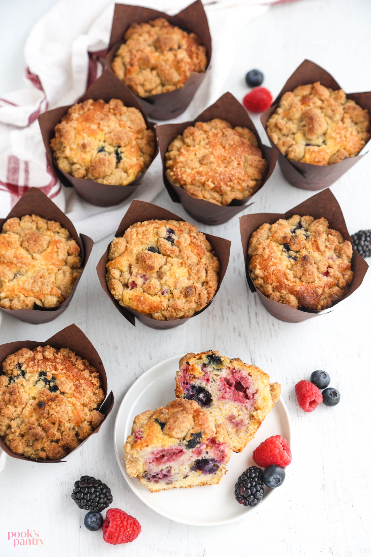 Bumbleberry muffins with crumble topping on a white wood background with berries scattered around.
There is a muffin sliced in half on a small white plate in the foreground.