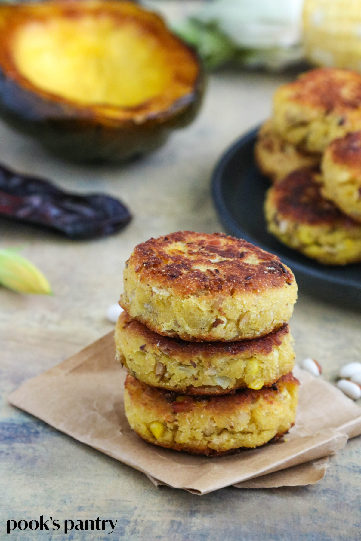 Three Sisters squash fritters stacked on top of each on a folded piece of brown parchment paper. There are fall vegetables in the background.