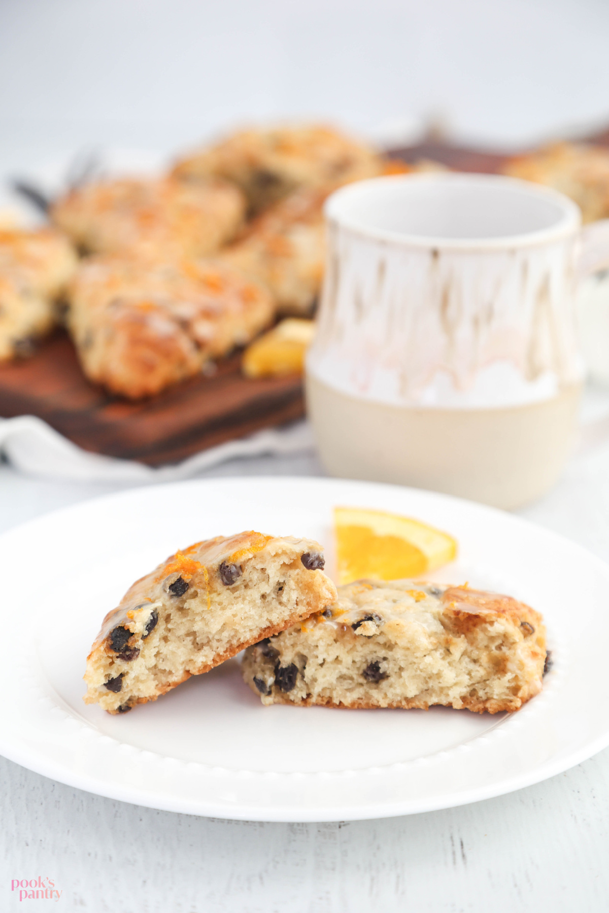 A scone, broken in half, on a small white plate, with a handmade ceramic mug behind it. There is a cutting board with scones on it in the background.
