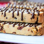 Butter pecan biscotti, drizzled with chocolate, on a white platter, sitting on top of a red tablecloth. The chocolate drizzle is on the side of the biscotti, flowing over the top.