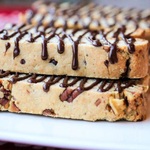 Butter pecan biscotti, drizzled with chocolate, on a white platter, sitting on top of a red tablecloth. The chocolate drizzle is on the side of the biscotti, flowing over the top.