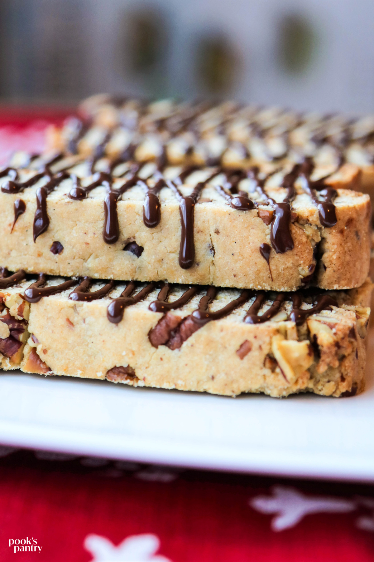 Butter pecan biscotti, drizzled with chocolate, on a white platter, sitting on top of a red tablecloth. The chocolate drizzle is on the side of the biscotti, flowing over the top.