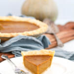 A slice of Hubbard squash pie on a small white plate with a silver fork on the side. There is a pie in the background, next to a Hubbard squash.