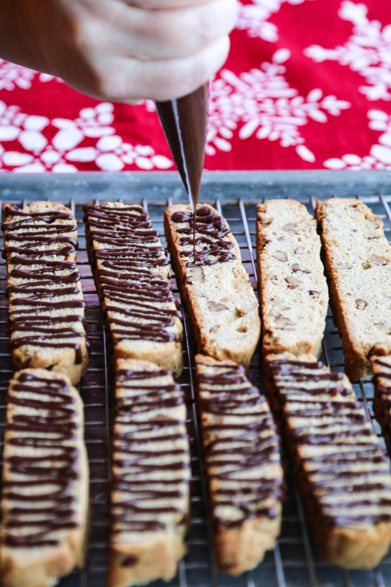 Drizzling chocolate, using a piping bag, onto butter pecan biscotti on a sheet pan.
