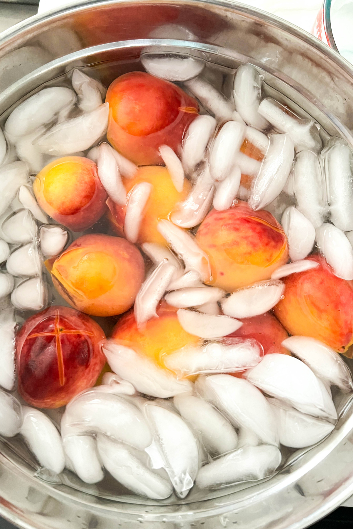 Peaches in a bowl of ice water, ready to be peeled. Each peach has an "x" cut into the bottom of it showing how to score them for peeling.