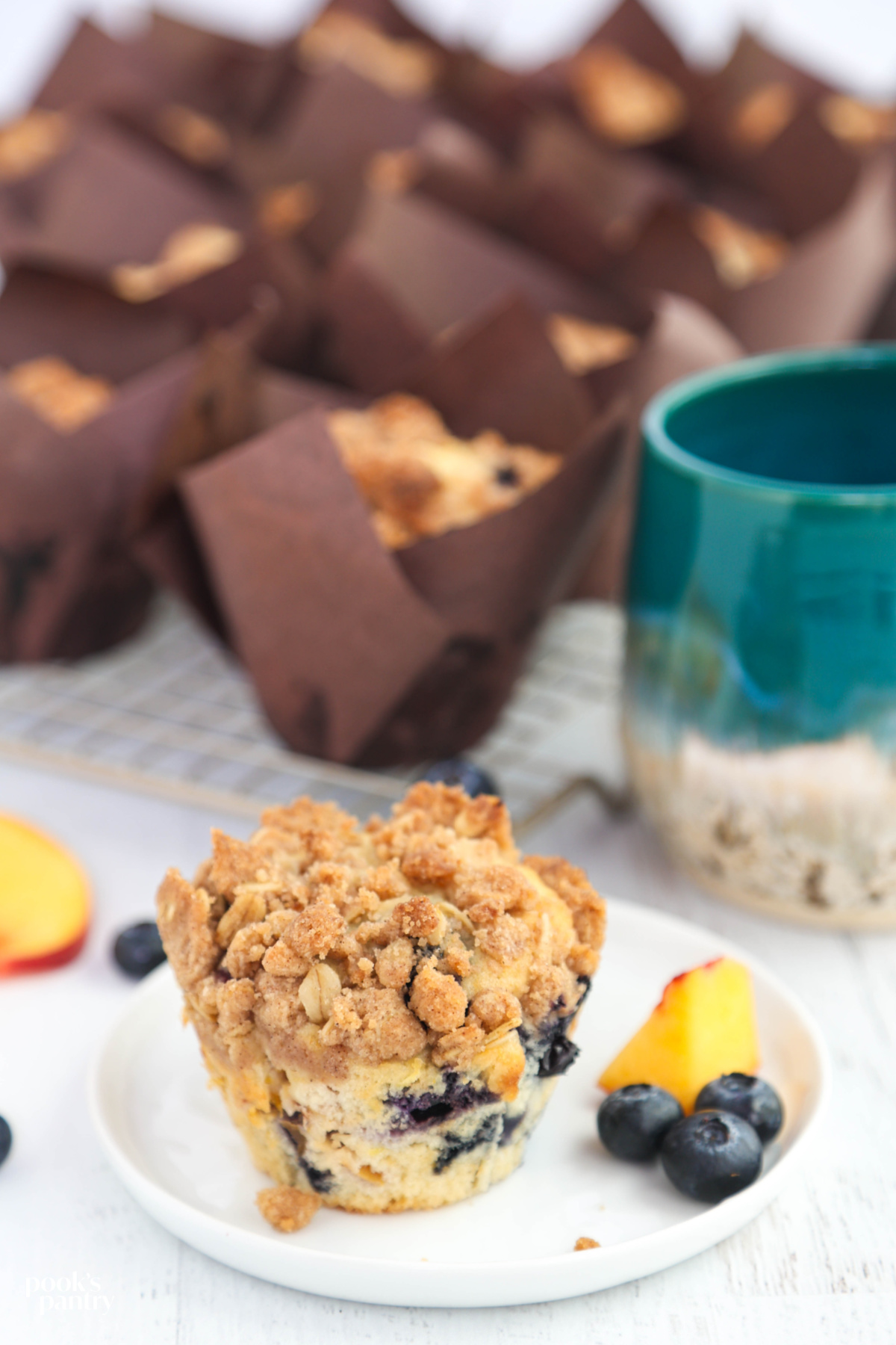 A crumb-topped muffin on a small white plate with a piece of peach and a few blueberries next to it.