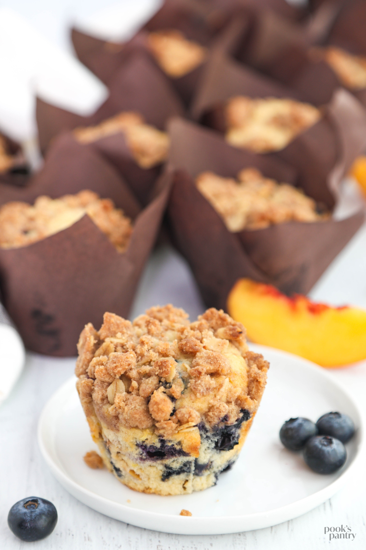 Crumb-topped fruit muffin with peaches and blueberries. There are more muffins in the background.